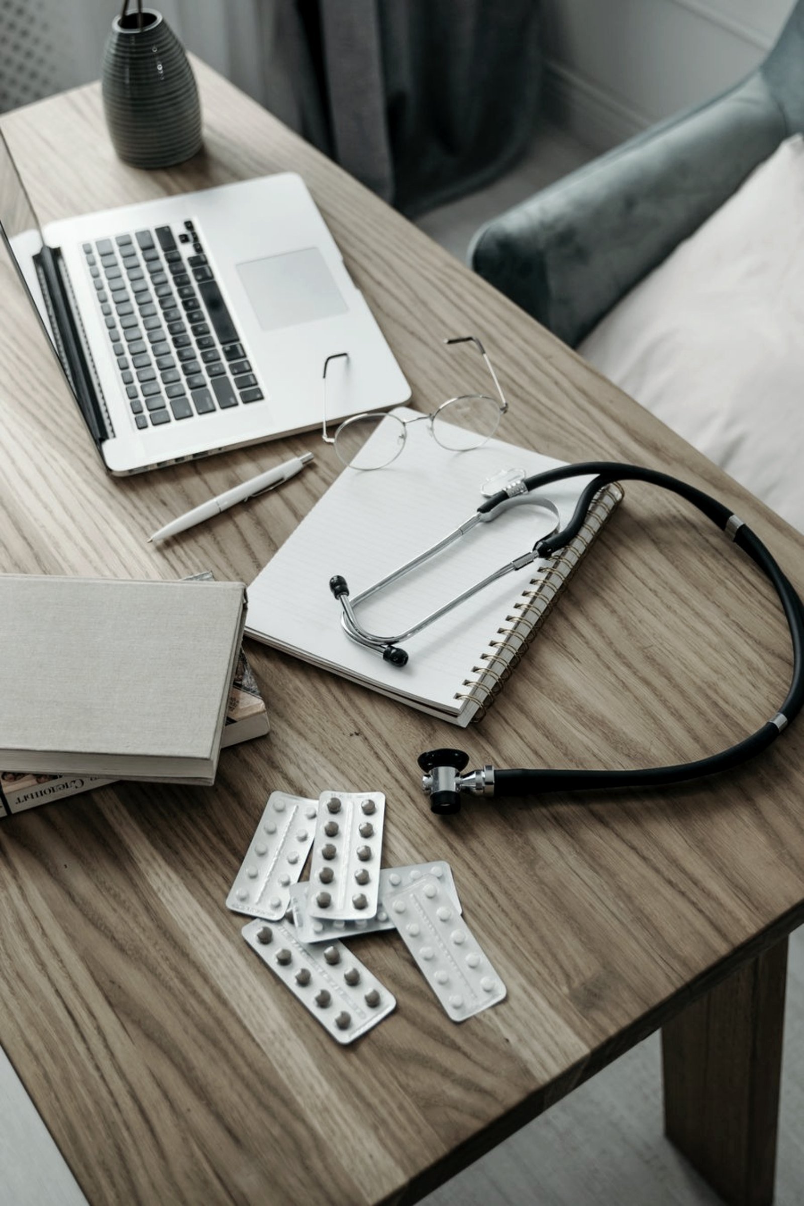 A desk with a laptop, notebook, reading glasses, blister packs and a stethoscope